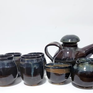 May include: A ceramic tea set featuring a dark brown teapot with a lid and handle, accompanied by four matching rounded cups and a small creamer pitcher. Each piece has a glossy finish with subtle blue and grey glaze drips along the rims.