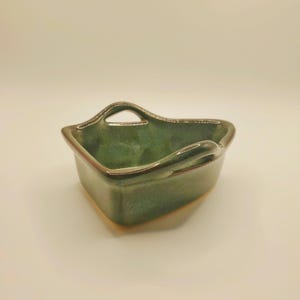 May include: A small, triangular ceramic bowl with a glossy, mottled sage green glaze. The bowl features two integrated handles on opposite sides, with a smooth, curved rim and a deep basin, showcasing a consistent, reflective finish throughout.
