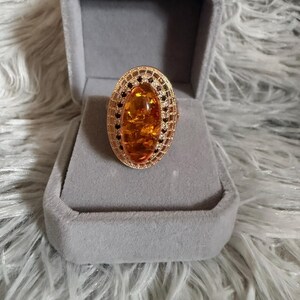 May include: An oval-shaped ring featuring a large, translucent amber-colored stone at the center. The stone is surrounded by a decorative gold-toned metal frame with intricate filigree detailing and small, dark accent stones embedded along the border.
