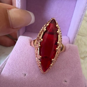 May include: A marquise-cut red gemstone ring set in a rose gold-toned metal band with intricate filigree detailing along the edges. The ring features a prominent, elongated, faceted red stone held by small prongs within a decorative metal setting.