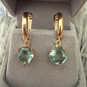 May include: A pair of rose gold hoop earrings featuring dangling, faceted, pentagonal light green gemstones. The metal hoops have a polished finish and a visible hallmark stamp on the inner side, securing the stones with small metal prongs.