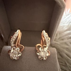 May include: A pair of rose gold earrings featuring a flower-shaped design with clear stones at the center and a curved, leaf-like accent set with small clear stones along the upper portion of the earring structure, presented in a gray box.