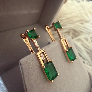 May include: A pair of rose gold drop earrings featuring a square-cut green gemstone at the top and a larger rectangular-cut green gemstone at the base, connected by a vertical bar accented with small clear stones in a hinged setting.