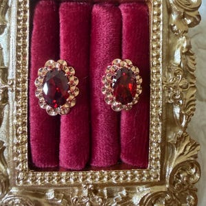 May include: A pair of oval-shaped stud earrings featuring deep red faceted gemstones surrounded by a scalloped gold-toned metal setting. The earrings are displayed on a dark red velvet insert within an ornate, rectangular gold-colored frame.