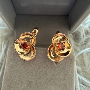 May include: A pair of gold-toned earrings featuring a swirling, layered metal design that resembles a flower. Each earring is centered with a single, round, faceted orange-red gemstone. The earrings have a polished finish and a secure lever-back closure.
