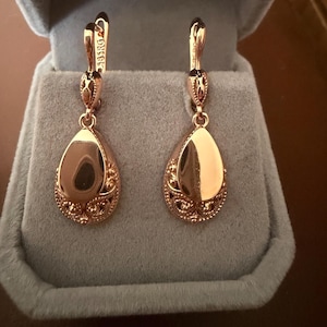 May include: A pair of rose gold-toned drop earrings featuring a teardrop-shaped pendant with a smooth, polished center and intricate filigree detailing at the base. The earrings have a hinged lever-back closure with the text 585RG engraved on the metal.