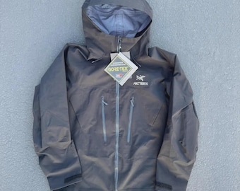 Puede incluir: Una chaqueta con capucha de color gris oscuro que presenta una cremallera frontal completa, dos bolsillos verticales con cremallera en el pecho y puños ajustables. Incluye el logotipo de Arc'teryx y una etiqueta de Gore-Tex.