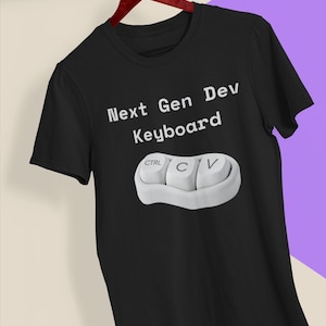 Op de afbeelding: Een zwart t-shirt met korte mouwen voorzien van de witte tekst Next Gen Dev Keyboard boven een afbeelding van drie witte computertoetsen met de labels CTRL, C en V. Het shirt heeft een ronde hals en hangt aan een rode kledinghanger.