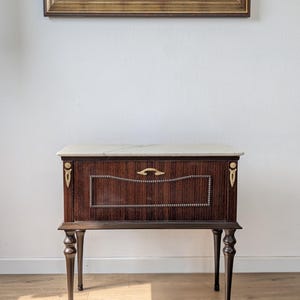 May include: A dark wood side table featuring a rectangular white marble top, a single drawer with a curved brass handle, and decorative brass accents on the upper corners. The table stands on four slender, tapered legs with a dark brown finish.