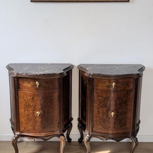 May include: A pair of wooden bedside cabinets featuring dark brown burl wood veneer, curved fronts, and grey marble tops. Each cabinet includes one drawer and one door with brass hardware, supported by four curved legs with carved detailing.