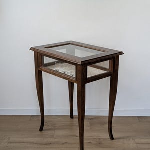 Può includere: Un tavolo espositore rettangolare in legno con piano in vetro e pannelli laterali in vetro. Il tavolo presenta quattro gambe curve e un ripiano interno di colore chiaro che ospita diversi piccoli bicchieri trasparenti all'interno.