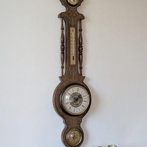 Peut inclure: Une station météo murale verticale avec un cadre en bois foncé orné de volutes dorées. Elle comprend un baromètre circulaire en haut, un thermomètre central à échelle verticale, une horloge à chiffres romains et une boussole en bas.