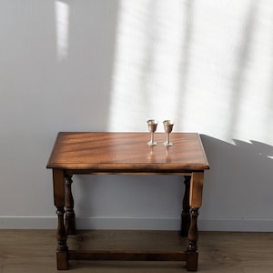 May include: A rectangular wooden side table with a smooth, polished brown surface and four turned legs connected by a lower stretcher. Two small, metallic silver-toned goblets with slender stems and rounded bowls sit on the tabletop.