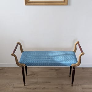 Puede incluir: Un banco con asiento rectangular tapizado en tela azul claro con un patrón repetitivo en azul oscuro. La estructura incluye apoyabrazos dorados curvos con detalles en madera oscura, sostenido por cuatro patas cónicas de color marrón.