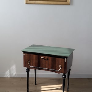 Puede incluir: Una mesa auxiliar rectangular con acabado de veta de madera marrón oscuro y una superficie superior de color verde salvia. La mesa cuenta con cuatro patas negras cónicas, un cajón con un tirador dorado horizontal y adornos metálicos dorados.