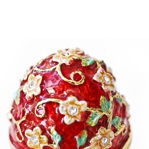 May include: A red decorative egg with a floral design. The egg features white flowers with crystal accents, gold-colored vines, and green leaves. The egg is a decorative item.