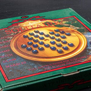 May include: A green box featuring a Solitaire board game. The game board is a gold-colored circle with blue marbles. The box has text that reads "A Traditional Wooden Board Game with Glass Marbles."