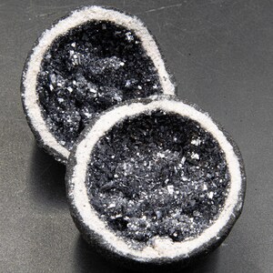 Puede incluir: Dos mitades de una geoda, que revelan un interior oscuro y cristalino con inclusiones brillantes de color plateado. El borde exterior es gris oscuro con un borde blanco, sobre un fondo gris oscuro. La geoda es un artículo decorativo natural.