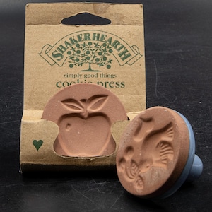 May include: A terracotta cookie press with a blue handle, featuring an apple design and a bird design. The packaging reads "SHAKERHEARTH simply good things cookie press."