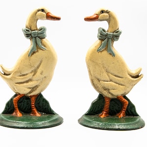 May include: A pair of duck ornaments. The ducks are painted cream with orange beaks and feet. Each has a light blue bow and stands on a green base. The figurines face each other, creating a symmetrical arrangement. The ornaments are likely made of cast iron.