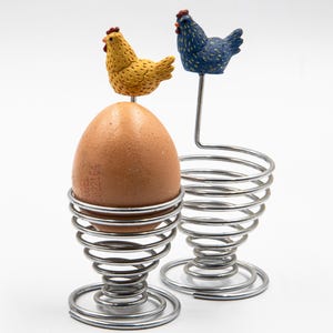 May include: Two egg cups with chicken ornaments. One cup holds a brown egg. The egg cups are made of coiled metal. One chicken is yellow, the other is blue. The chickens have red combs.