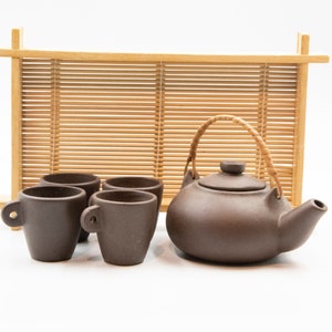 Op de afbeelding: Een bruine keramische theeset met een theepot, vier kleine kopjes en een bamboe scherm. De theepot heeft een gevlochten handvat en een tuit. De kopjes hebben kleine handvatten. De set is op een wit oppervlak geplaatst.