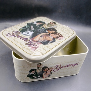 May include: A rectangular metal tin with a hinged lid, featuring a romantic vintage design. The lid and sides display images of couples and the word "Greetings" in a decorative font. The tin is off-white with a gold trim.