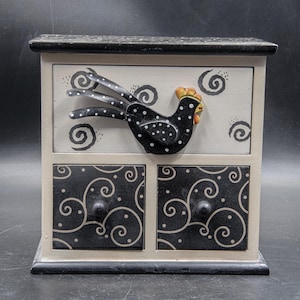 May include: A decorative wooden storage box with two drawers. The box is painted in cream, black, and white. A black and white polka-dotted rooster is affixed to the front. The drawers have black swirl designs and black knobs.