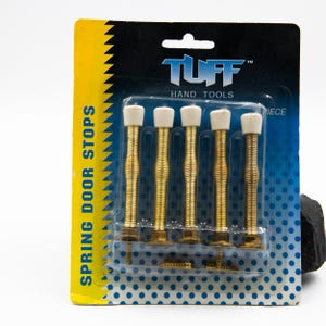 May include: A pack of five spring door stops. Each door stop features a gold-coloured, coiled spring with a white rubber tip. The packaging is blue with white polka dots and the words "SPRING DOOR STOPS" in yellow.