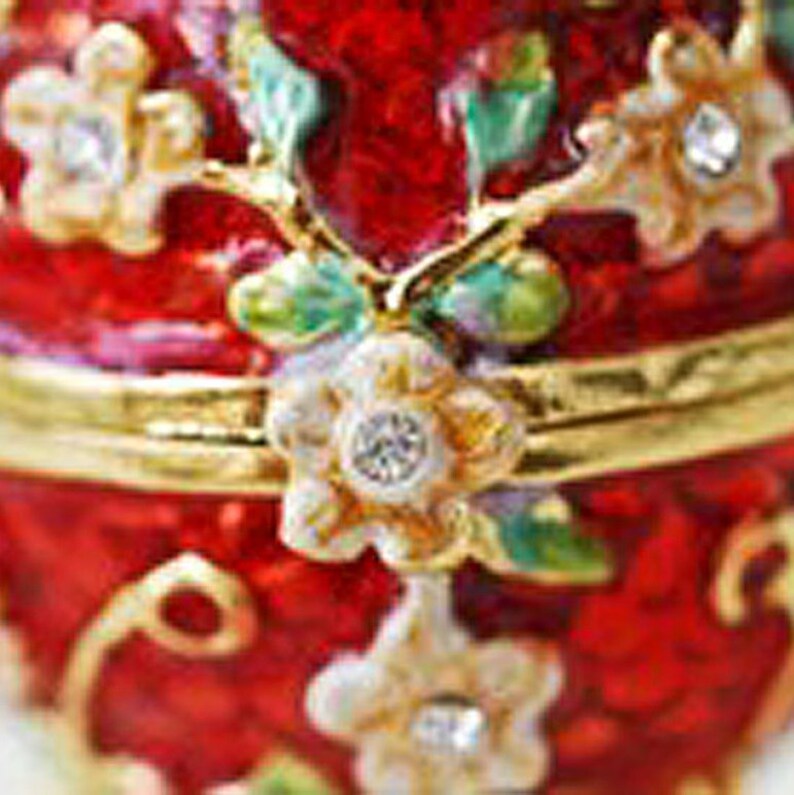 May include: A red, ornate trinket box with gold accents and floral embellishments. The box features a hinged lid and is adorned with white and gold flowers with crystal centers. Green and purple leaves accent the flowers.