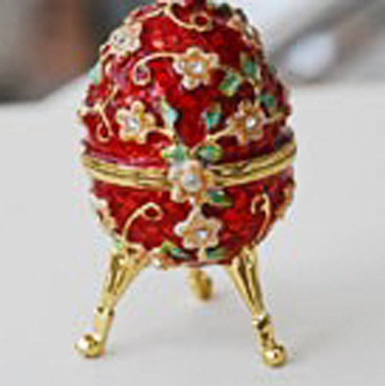 May include: A red enameled egg-shaped trinket box with gold accents and floral detailing. The box is supported by three gold-colored legs. The egg is decorated with white and clear gemstones.