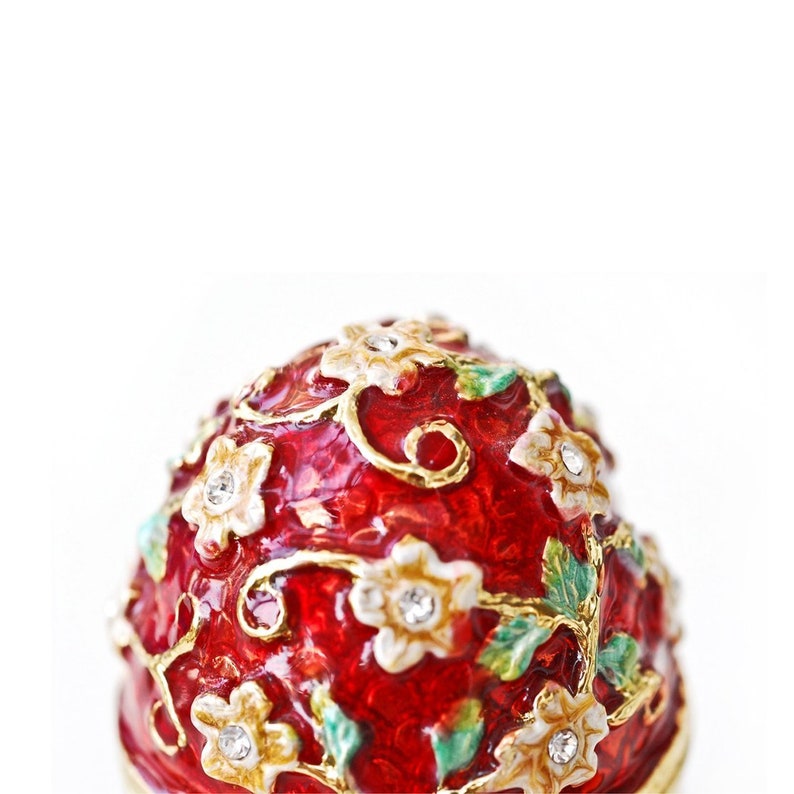 May include: A red decorative egg with a floral design. The egg features white flowers with crystal accents, gold-colored vines, and green leaves. The egg is a decorative item.