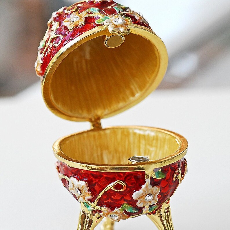 May include: A decorative, open egg-shaped trinket box. The red enamel exterior is adorned with white floral accents and gold trim. The interior is gold-colored. The box is supported by three gold legs and has a magnetic closure.