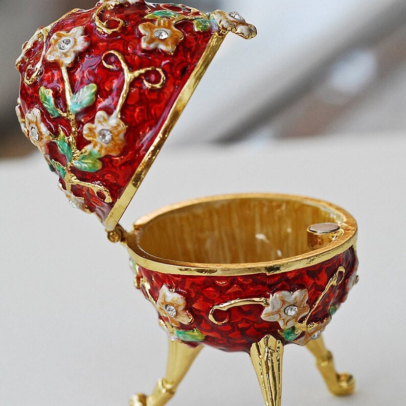 May include: A red, decorative egg-shaped trinket box with a hinged lid. The box is adorned with white and gold floral designs and small, sparkling embellishments. The interior is gold-colored, and the box rests on three gold legs.
