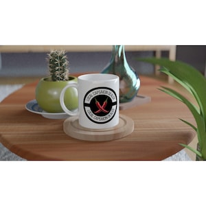 Può includere: Una tazza in ceramica bianca con una grafica circolare nera raffigurante due peperoncini rossi incrociati e il testo LOCAL CAPSAICIN DEALER stampato due volte lungo il bordo. La tazza è dotata di un manico e poggia su un sottobicchiere in legno.