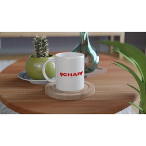 Può includere: Una tazza in ceramica bianca con la parola SCHARF stampata in lettere maiuscole rosse con un effetto gocciolante. La tazza ha un manico standard e poggia su un sottobicchiere circolare in legno, posizionato su un tavolo rotondo.