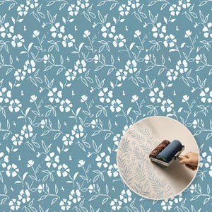 Pode incluir: Um padrão de flores e folhas brancas desenhadas à mão sobre um fundo azul poeirento. Uma inserção circular mostra um rolo de pintura texturizado aplicando uma estampa botânica de samambaia em azul claro sobre uma superfície esbranquiçada.