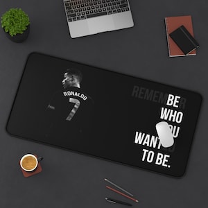 May include: A rectangular black desk mat featuring a monochrome graphic of a soccer player wearing a jersey with the name RONALDO and the number 7 on the back. The text REMEMBER BE WHO YOU WANTED TO BE is printed in white on the right side.
