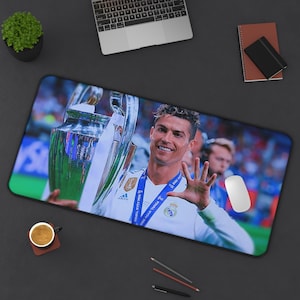 May include: A rectangular desk mat featuring a high-resolution print of a soccer player in a white jersey holding a large silver trophy. The player is smiling and holding up five fingers. The background shows a blurred stadium crowd in blue tones.