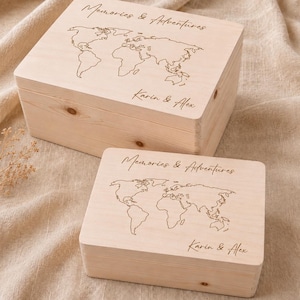 Puede incluir: Dos cajas rectangulares de madera clara con tapas abatibles. Cada caja presenta un mapa del mundo grabado con láser, el texto "Memories & Adventures" en la parte superior y los nombres "Karin & Alex" grabados en la esquina inferior derecha.