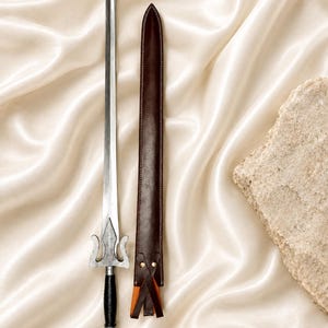 Può includere: Una spada in metallo argentato con lama appuntita, guardia decorativa e impugnatura nera, accompagnata da un fodero in pelle marrone scuro con cuciture visibili e rivetti in metallo. Il testo TheBladeSmithCrafts è stampato nell'angolo.