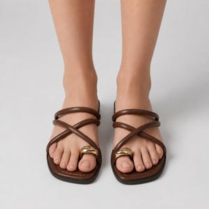 May include: A pair of dark brown leather slide sandals featuring two thin, rounded crossover straps across the top and a prominent, smooth gold-toned metal ring accent encircling the big toe on each sandal, set on a flat, square-toed sole.