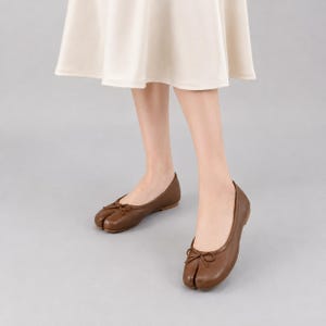 Könnte beinhalten: Ein Paar braune Leder-Ballerinas mit geteilter Zehenpartie und einer kleinen dekorativen Schleife auf der Vorderseite. Die Schuhe haben ein flaches Design mit abgerundeter Ferse und einer deutlichen Spalte zwischen dem großen Zeh und den anderen Zehen.