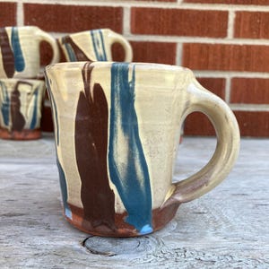 May include: A ceramic mug with a cream-colored base featuring vertical streaks of dark brown and blue glaze. The mug has a thick, curved handle and a reddish-brown base. The surface has a smooth, glossy finish with visible variations in the glaze.