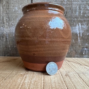Può includere: Un vaso rotondo in ceramica marrone fatto a mano con un'ampia apertura. Il vaso ha una finitura lucida e una combinazione di colori a strati, che passa da una parte superiore marrone scuro a una sezione centrale marrone più chiara e una base marrone rossiccio. Una moneta è accanto al vaso.