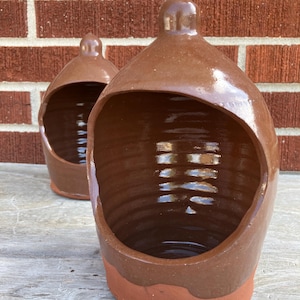 Puede incluir: Dos saleros de cerámica marrón con un acabado brillante en la parte superior y una base mate sin esmaltar. Cada recipiente presenta una abertura ancha y arqueada en la parte frontal y un pequeño pomo en la parte superior, con una textura interior estriada.