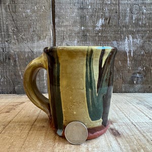 Może przedstawiać: Ręcznie robiony ceramiczny kubek z beżową podstawą i brązowym uchwytem. Kubek ma pionowe pociągnięcia pędzla w kolorze zielonym i brązowym, z odrobiną niebieskiego na krawędzi. Obok kubka znajduje się moneta jako odniesienie do rozmiaru.