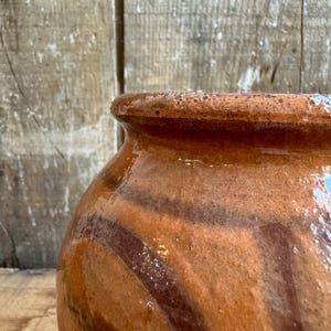 May include: Close-up of a rustic, glazed ceramic pot. The pot features a warm, earthy color palette with shades of brown and orange, and dark brown stripes. The rim is slightly flared, and the surface has a glossy finish, suggesting it's been fired.