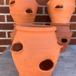 May include: A collection of terracotta strawberry pots. Each pot has a round opening and several side openings for planting. The pots are a warm, earthy orange color and are stacked on a wooden surface.