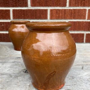 May include: Two small, round, brown glazed ceramic pots with a smooth, shiny finish and a slightly flared rim. Each pot features a subtle, horizontal decorative band of small, repeating indentations around the upper body of the vessel.